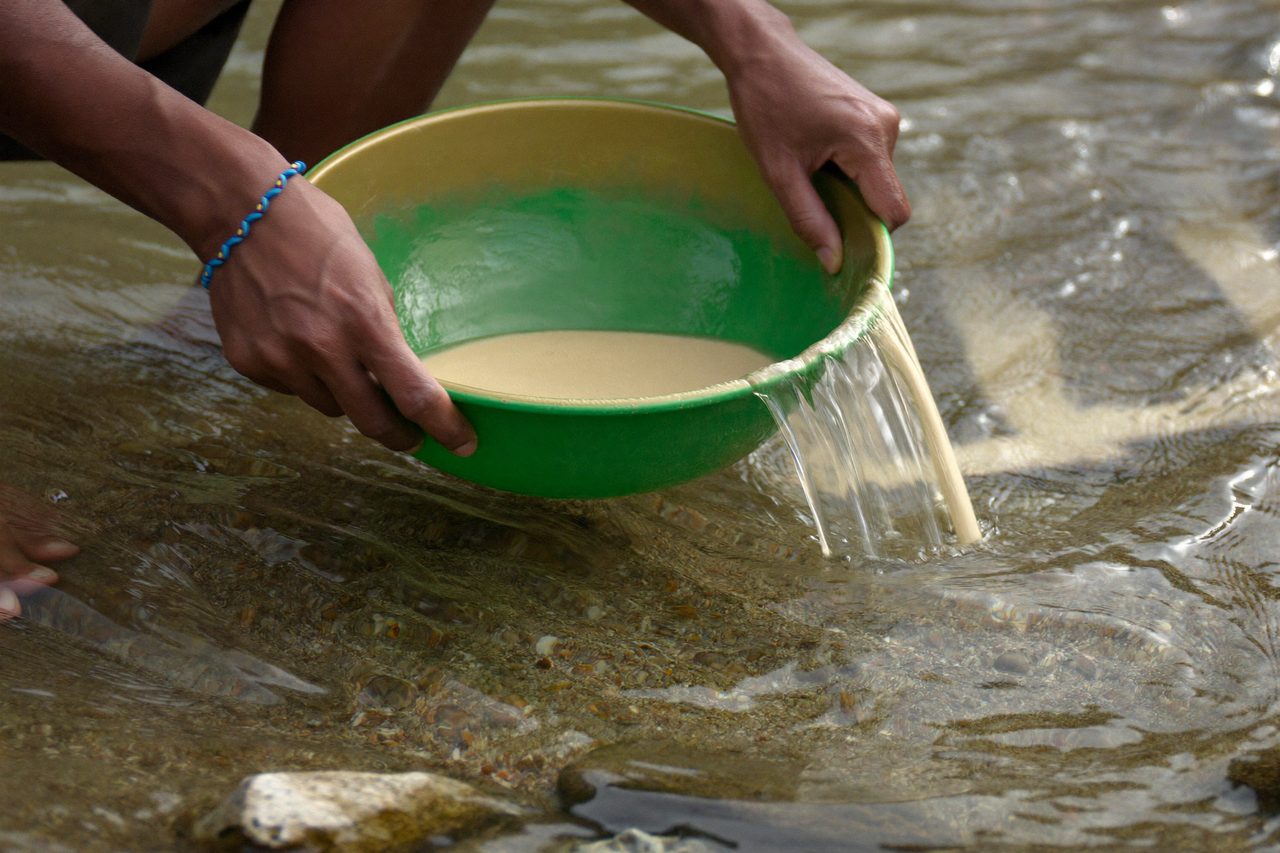 Two hands swirling the pan under water, sand washing over the rim